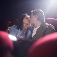 Low angle portrait of young couple kissing in cinema while enjoying romantic date, man embracing girlfriend in dark hall, copy space